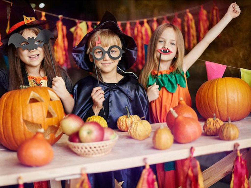 سه کودک با گریم و لباس مخصوص جشن با میز جلوی آن‌ها که با کدو تنبل و سیب به سبک دکور هالووین آراسته شده است.