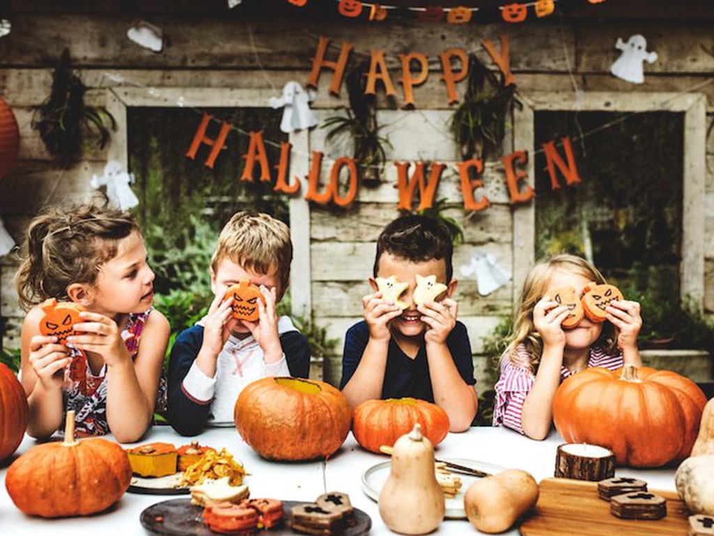 کودک که با شیرینی‌های مخصوص هالووین صورت خود را پوشش داده‌اند و میزی که با خوراکی‌های مخصوص و کدو تنبل چیدمان شده است.