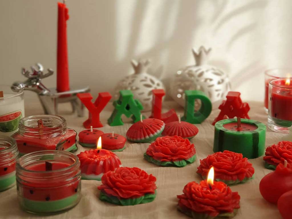 شمع به شکل حروف یلدا با ترکیب‌بندی رنگ‌های سبز و قرمز در کنار شمع‌هایی به شکل گل و هندوانه خودنمایی می‌کنند.