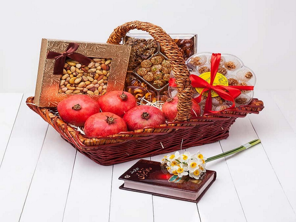 سبد حصیری پر شده با میوه، آجیل و انواع شکلات در کنار گل نرگس و دیوان حافظ هدیه خوبی برای شب یلدا است.