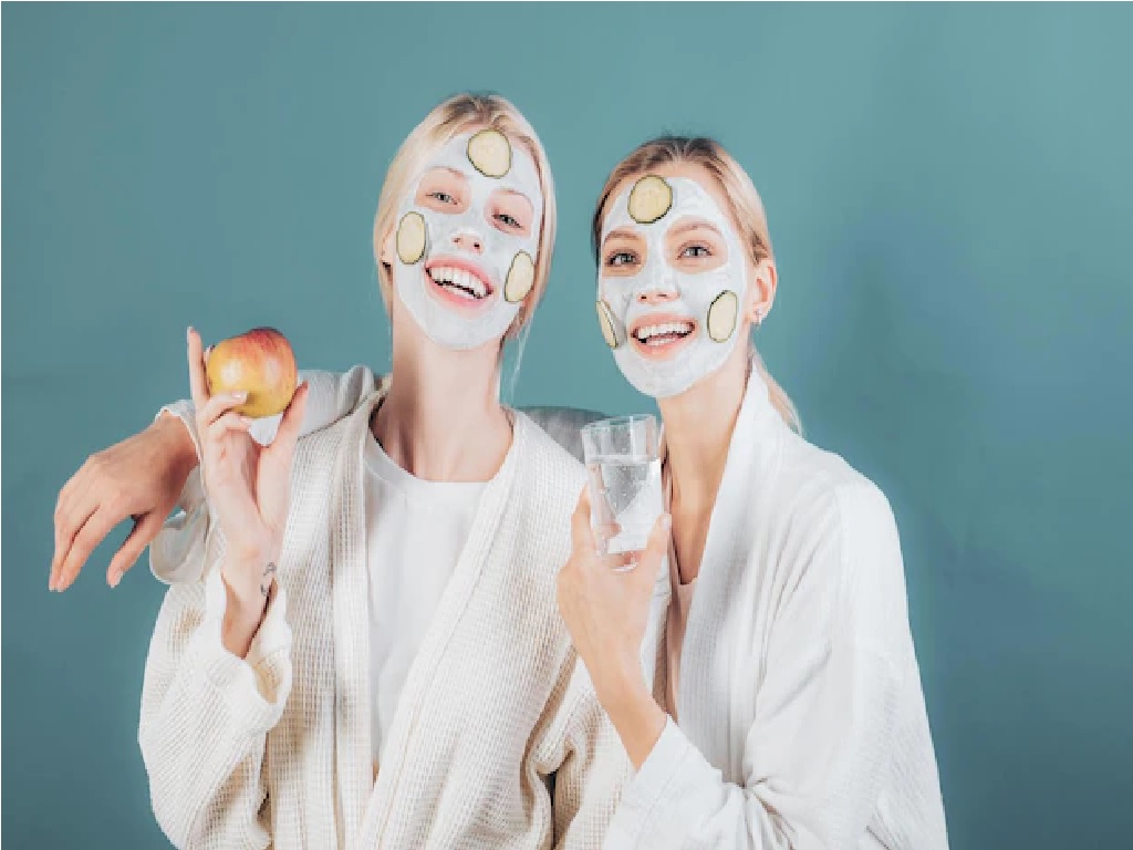 دو دختر جوان با استفاده از ماسک لبنیات و نشان دادن آب و سیب به عوامل سلامتی پوست اشاره می‌کنند.